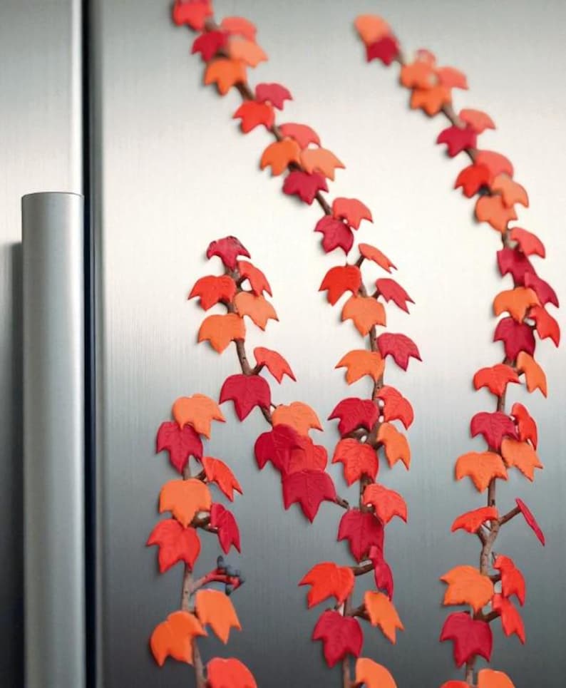 May include: Decorative fall-themed leaf vines in shades of red and orange. The artificial leaves are attached to brown stems, creating a realistic autumn aesthetic. The vines are arranged vertically against a metallic background.