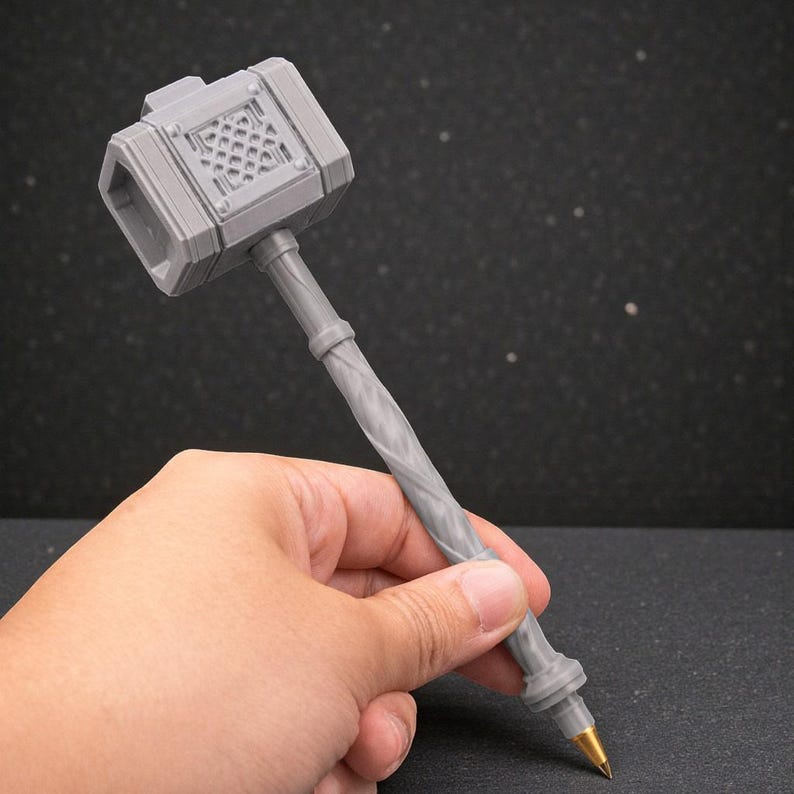 May include: A grey pen in the shape of a hammer, held in a hand. The hammer head has a detailed design, and the handle is twisted. The pen tip is gold-coloured. The background is dark with small white dots.