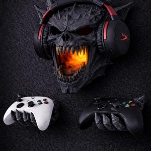 May include: A dark gray demonic skull with red-accented headphones and a fiery glow emanating from its mouth. Below, two game controllers, one white and one black, are held by clawed hands, suggesting a gaming theme.