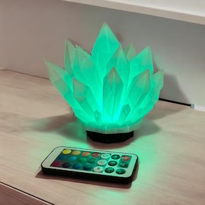 May include: A glowing, green crystal-shaped lamp with a black base. The lamp is lit from within, and a remote control with color options sits in front of it. The lamp is on a wooden surface.