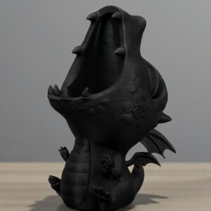 May include: Black 3D-printed dragon figurine with an open mouth, small wings, and detailed scales. The dragon is sitting with its mouth open, ready to hold small items. The figurine is on a wooden surface.