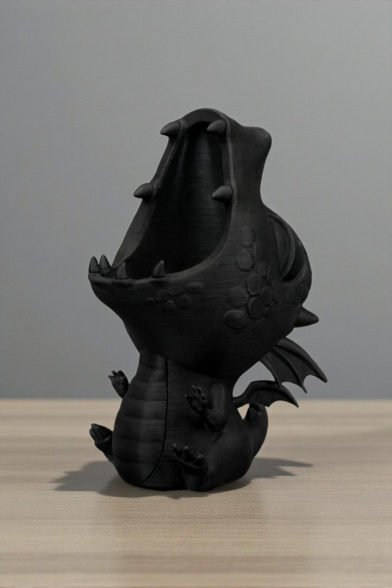 May include: Black 3D-printed dragon figurine with an open mouth, small wings, and detailed scales. The dragon is sitting with its mouth open, ready to hold small items. The figurine is on a wooden surface.