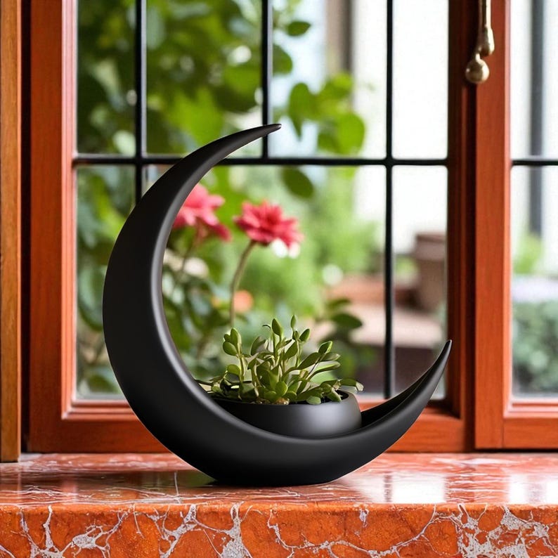 May include: A black ceramic crescent moon-shaped planter holding a small potted plant. The planter sits on a red and orange marble surface in front of a window with a view of green foliage and red flowers.