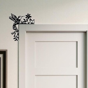 May include: Black metal hummingbird and floral corner decor. The hummingbird is depicted in flight, with detailed wing and feather designs. The flowers and leaves are also intricately cut, creating a decorative accent for a door or wall.