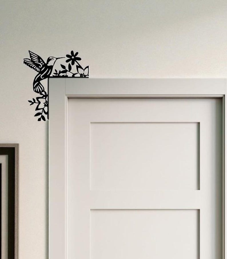 May include: Black metal hummingbird and floral corner decor. The hummingbird is depicted in flight, with detailed wing and feather designs. The flowers and leaves are also intricately cut, creating a decorative accent for a door or wall.