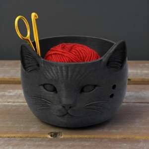 May include: A black cat-shaped yarn bowl containing a red ball of yarn. The bowl features a detailed cat face design, complete with ears and whiskers. Gold scissors are placed on the side. The bowl rests on a wooden surface.