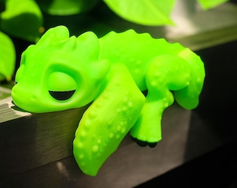May include: A bright green, stylized lizard figurine with a smooth body and textured legs. The figurine is resting on a dark surface, with green leaves in the background. The lizard has a closed eye and a smiling expression.
