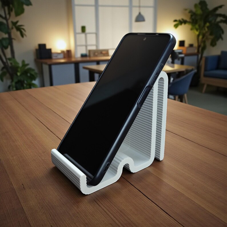 Op de afbeelding: Een witte, geribbelde telefoonstandaard met een zwarte smartphone. De standaard heeft een gebogen ontwerp en is gemaakt van een stevig materiaal. De telefoon is schuin geplaatst voor gemakkelijk kijken. De standaard staat op een houten tafel.
