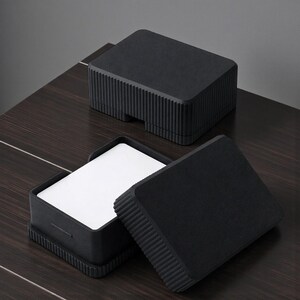 May include: Black, textured box with a lid, containing white cards. The box has a grooved design on the sides and is open, revealing the cards inside. Another closed box is in the background.