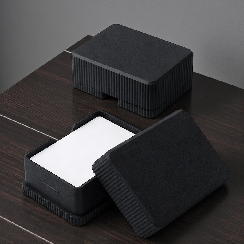 May include: Black, textured box with a lid, containing white cards. The box has a grooved design on the sides and is open, revealing the cards inside. Another closed box is in the background.
