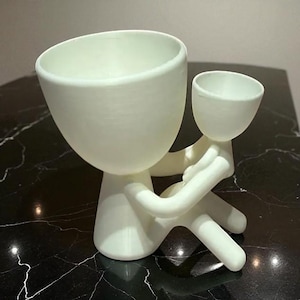 Op de afbeelding: Een witte, gestileerde plantenbak in de vorm van een persoon die een kleinere plantenbak vasthoudt. De grotere plantenbak heeft een afgeronde, open bovenkant en de kleinere wordt in de armen van de grotere figuur gehouden. Het ontwerp is minimalistisch en modern.
