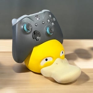 May include: A yellow Psyduck figure with a grey Xbox controller resting on top. The controller has blue accents and the Xbox logo. The figure has a light beige beak and white eyes. The background is a light brown wooden surface.