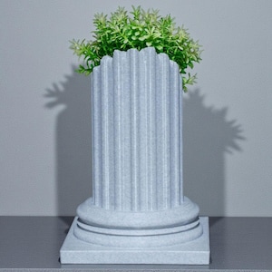 May include: A light grey, fluted column-shaped planter with a square base, holding a vibrant green plant. The planter has a classic architectural design, suitable for indoor or outdoor use. The plant adds a natural touch to the decorative piece.