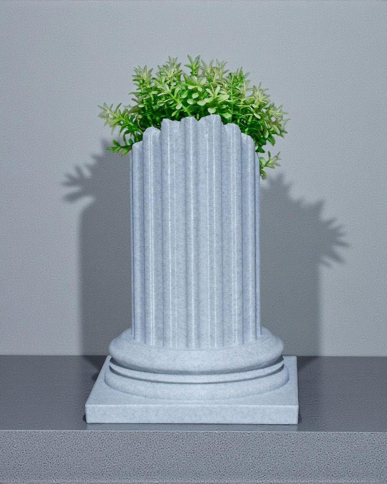 May include: A light grey, fluted column-shaped planter with a square base, holding a vibrant green plant. The planter has a classic architectural design, suitable for indoor or outdoor use. The plant adds a natural touch to the decorative piece.