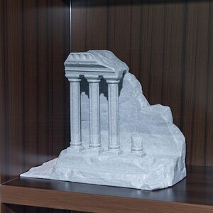 May include: A gray stone sculpture of a classical temple ruin with three columns. The sculpture has a weathered appearance and sits on a flat base. The background is a dark wood surface.