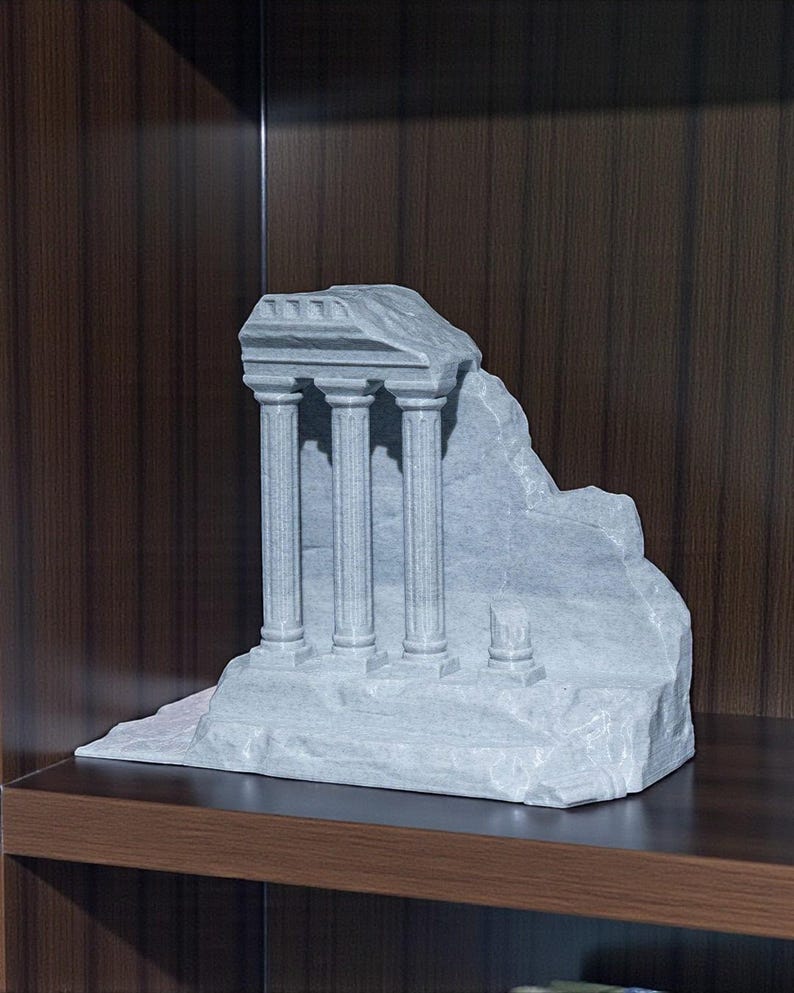 May include: A gray stone sculpture of a classical temple ruin with three columns. The sculpture has a weathered appearance and sits on a flat base. The background is a dark wood surface.