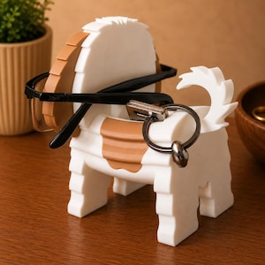 May include: A white and brown dog-shaped eyeglasses and key holder. The holder has a round head and a tail. Black eyeglasses and a key ring are placed on the holder. The holder is on a wooden surface.