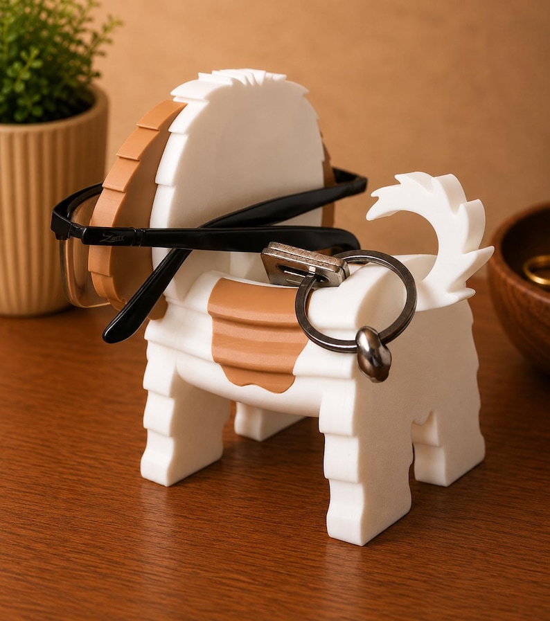 May include: A white and brown dog-shaped eyeglasses and key holder. The holder has a round head and a tail. Black eyeglasses and a key ring are placed on the holder. The holder is on a wooden surface.