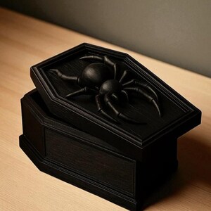 May include: A black, coffin-shaped box with a hinged lid. The lid features a raised relief of a large spider. The box is made of wood and has a matte finish. The box is open, revealing the interior.