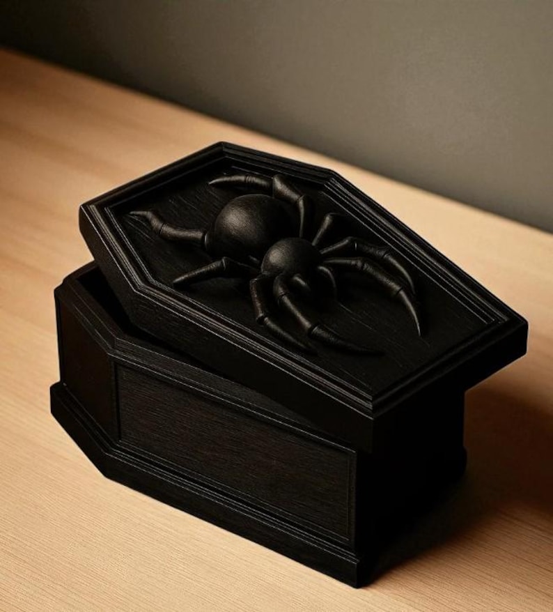 May include: A black, coffin-shaped box with a hinged lid. The lid features a raised relief of a large spider. The box is made of wood and has a matte finish. The box is open, revealing the interior.
