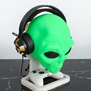 May include: A green alien head-shaped headphone stand with a white base, holding a white game controller. Black headphones with gold accents rest on the head. The stand is on a black marble surface.