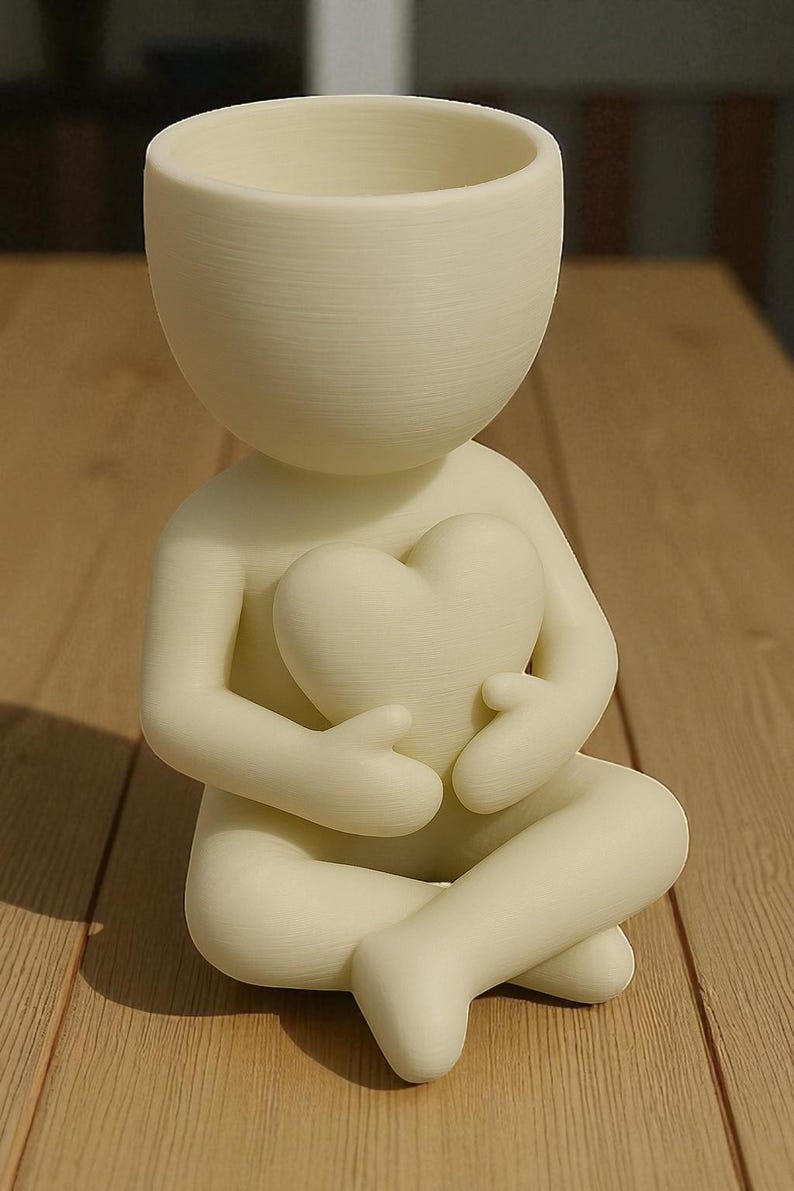 May include: A cream-colored figurine of a person holding a heart, with a planter-like head. The figure is seated with legs crossed. The heart is held in the figure's lap. The figurine is made of a smooth material and sits on a wooden surface.