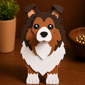 May include: A layered papercraft sculpture of a Shetland Sheepdog, featuring brown, white, and black colors. The dog has large, expressive eyes and a fluffy mane. The artwork is displayed on a wooden surface.