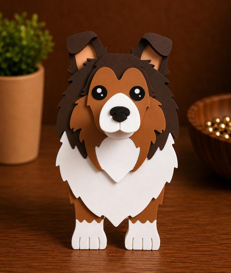 May include: A layered papercraft sculpture of a Shetland Sheepdog, featuring brown, white, and black colors. The dog has large, expressive eyes and a fluffy mane. The artwork is displayed on a wooden surface.