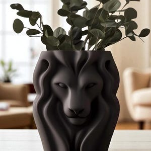 May include: A dark grey ceramic vase featuring a lion's face design, filled with green leafy branches. The lion's face is sculpted with detailed features. This decorative vase is a home accent, suitable for displaying flowers or foliage.