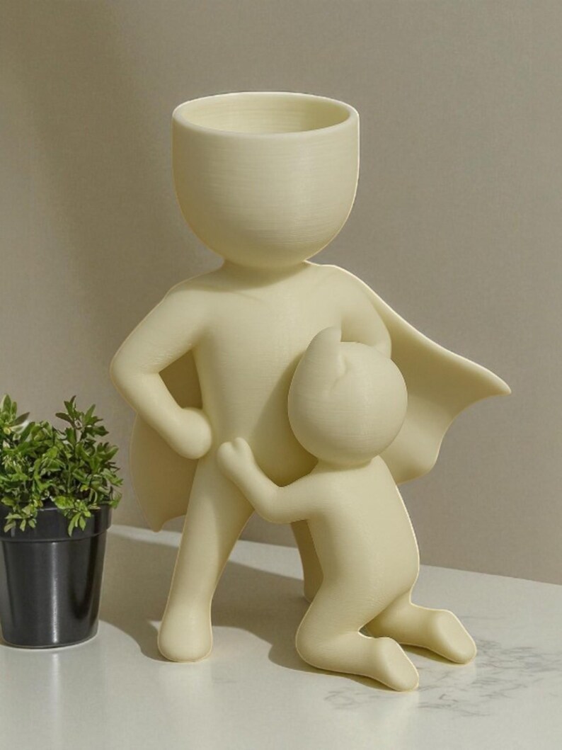 Op de afbeelding: Een cr&egrave;mekleurige keramische plantenbak in de vorm van een superheld met een kind dat zich aan zijn been vastklampt. De superheld heeft een cape en een grote kop die dient als opening van de plantenbak. Een kleine potplant is zichtbaar op de achtergrond.