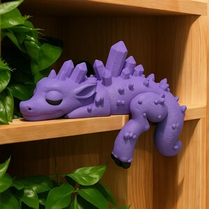 May include: A lavender-colored dragon figurine with crystal-like spikes along its back and head. The dragon is lying down with its eyes closed, and its tail curls over the edge of a wooden shelf. The dragon's feet are black.