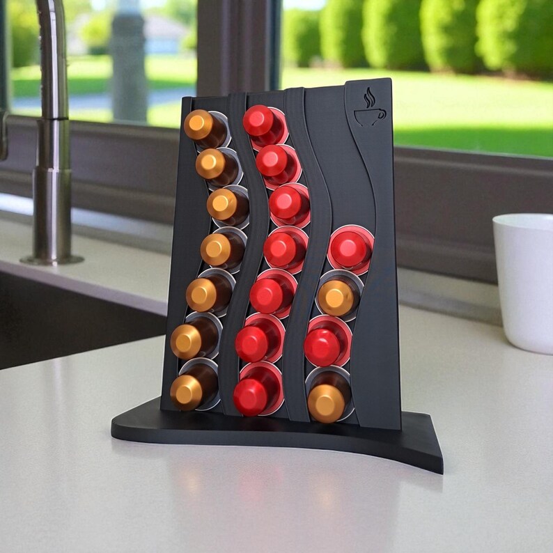 May include: A black coffee pod holder with a wave design, holding various coffee pods in gold and red. The holder has a coffee cup icon and is displayed on a white countertop near a window.