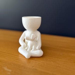Puede incluir: Una figura de cer&aacute;mica blanca de una persona sentada con un perro en su regazo. La figura tiene la cabeza ahuecada, lo que sugiere que podr&iacute;a usarse como una maceta peque&ntilde;a o un art&iacute;culo decorativo. La figura est&aacute; sobre una superficie de madera.