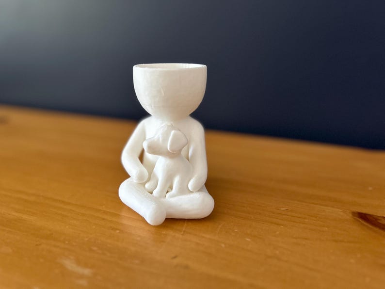 Puede incluir: Una figura de cer&aacute;mica blanca de una persona sentada con un perro en su regazo. La figura tiene la cabeza ahuecada, lo que sugiere que podr&iacute;a usarse como una maceta peque&ntilde;a o un art&iacute;culo decorativo. La figura est&aacute; sobre una superficie de madera.