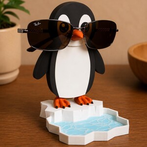 May include: A black, white, and orange penguin-shaped eyeglass holder with sunglasses. The penguin is standing on a white iceberg with a blue water design. The sunglasses are dark-lensed with a silver frame.