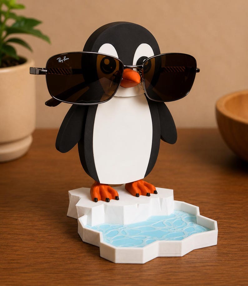 May include: A black, white, and orange penguin-shaped eyeglass holder with sunglasses. The penguin is standing on a white iceberg with a blue water design. The sunglasses are dark-lensed with a silver frame.