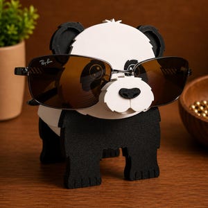 May include: A black and white panda-shaped eyeglass holder with a pair of brown-tinted sunglasses. The sunglasses have a black frame and the Ray-Ban logo. The holder is made of wood and has a cute, cartoonish design.