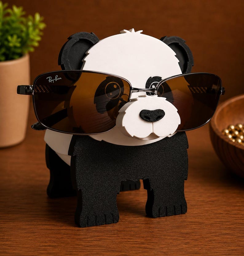 May include: A black and white panda-shaped eyeglass holder with a pair of brown-tinted sunglasses. The sunglasses have a black frame and the Ray-Ban logo. The holder is made of wood and has a cute, cartoonish design.