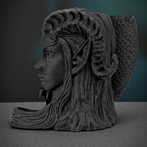 May include: A dark gray sculpture of a female figure with long hair, pointed ears, and horns. The sculpture has a textured finish and a detailed design, with a dragon-like tail on the back. The figure is in profile, with a serious expression.