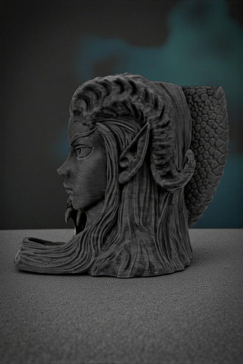 May include: A dark gray sculpture of a female figure with long hair, pointed ears, and horns. The sculpture has a textured finish and a detailed design, with a dragon-like tail on the back. The figure is in profile, with a serious expression.