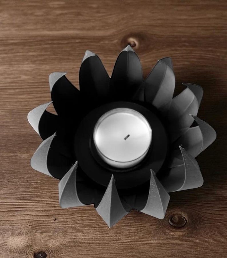 May include: Black metal candle holder in a flower shape with a white candle in the center. The petals have a folded design, and the holder sits on a wooden surface. A decorative accent for any room.