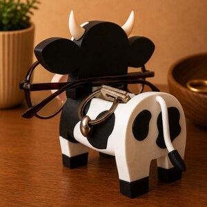 May include: A whimsical cow-shaped eyeglasses and key holder. The black and white painted wooden figurine has a tail, horns, and holds eyeglasses and keys. The cow is on a wooden surface.