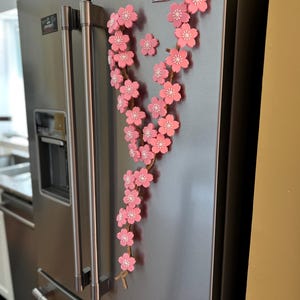 May include: A decorative pink cherry blossom branch with white accents, attached to a stainless steel refrigerator. The floral arrangement is made of multiple flowers and is a spring-themed home decor item.