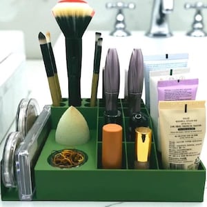 May include: A green makeup organizer filled with various beauty products. The organizer holds makeup brushes, mascara, lipstick, a makeup sponge, and other cosmetic items. The background includes a white countertop and a chrome faucet.