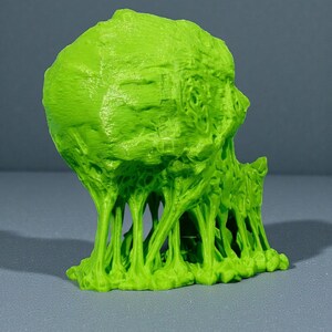 May include: A bright green, 3D-printed sculpture of a grotesque, alien-like head with a textured surface. The sculpture has a melting, dripping effect, with tendrils extending downwards. The object is set against a gray background.