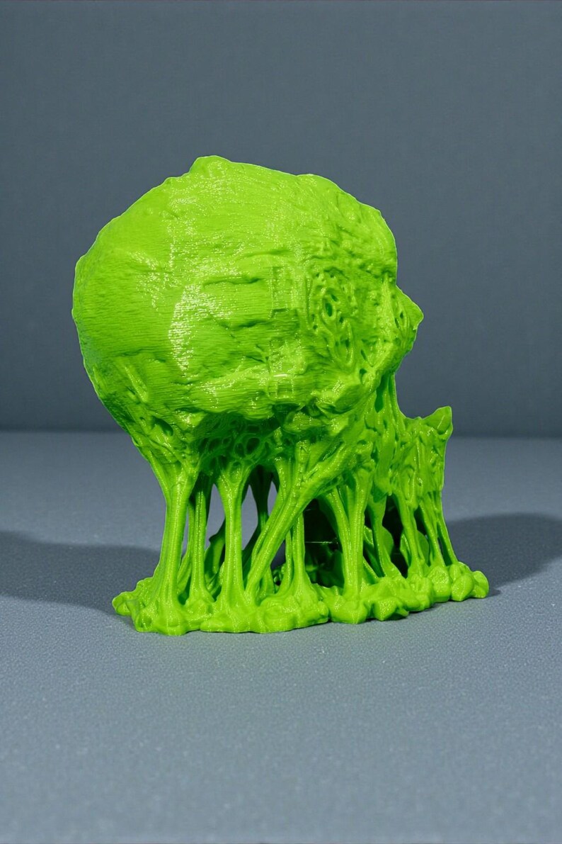May include: A bright green, 3D-printed sculpture of a grotesque, alien-like head with a textured surface. The sculpture has a melting, dripping effect, with tendrils extending downwards. The object is set against a gray background.