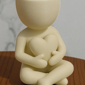 May include: A cream-colored figurine of a person holding a heart. The figure is seated with legs crossed and arms wrapped around the heart. The head of the figure is a small, open bowl shape. The figurine is made of a smooth, matte material.