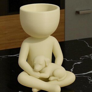 May include: A cream-colored figurine of a person seated with legs crossed, cradling a sleeping puppy. The figure has a large, open head, designed to hold small plants or items. The figurine is on a black, marble-like surface.