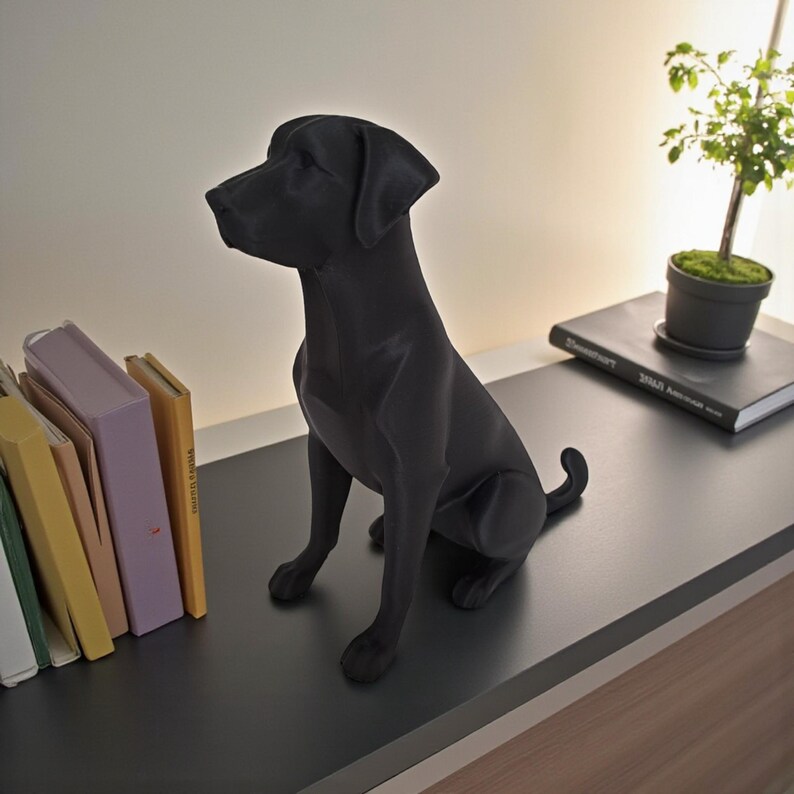 K&ouml;nnte beinhalten: Eine mattschwarze Labrador Retriever-Statue sitzt auf einer dunkelgrauen Oberfl&auml;che. Der Hund befindet sich in sitzender Position, mit eingerollter Rute. Eine kleine Topfpflanze und B&uuml;cher befinden sich im Hintergrund. Die Statue ist ein Wohnaccessoire.