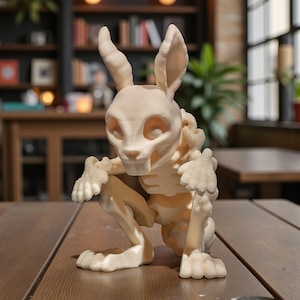 Puede incluir: Escultura de esqueleto de conejo beige, en posici&oacute;n agachada. La escultura presenta una estructura &oacute;sea detallada y orejas largas, expuesta sobre una mesa de madera. El fondo es un interior borroso con estanter&iacute;as y luces colgantes.